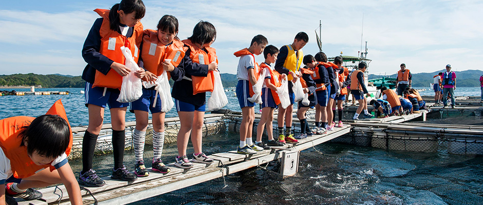 養殖されている鯛に餌やりをしている学生を写した写真。